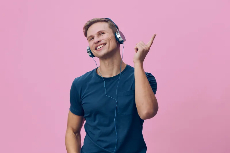 Joyful young man wearing headphones enjoying music with bright smile, pointing upward, casual blue t-shirt, light blue background, studio portrait, lifestyle concept, happy emotion, musical entertainm