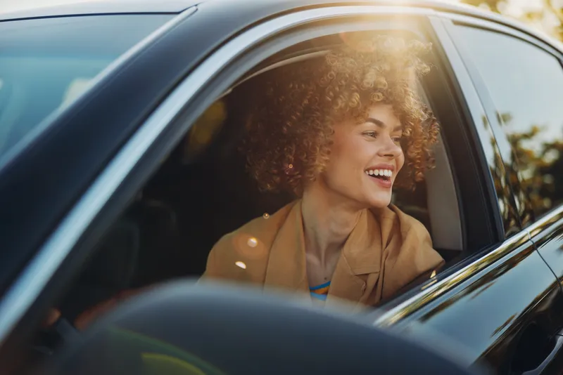 A joyful woman sits in a car, smiling from the passenger window as daylight glints on the glass, conveying a carefree lifestyle and retro film color aesthetic, warm and nostalgic, bright and candid.