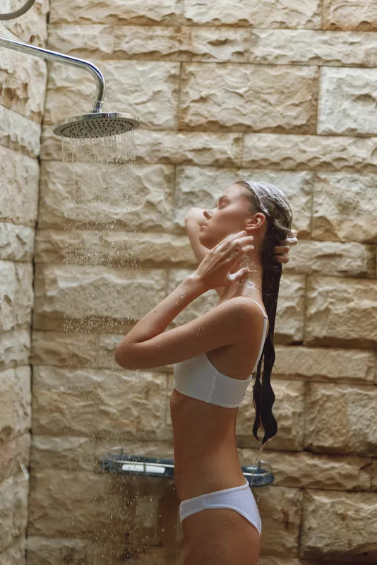 young woman showering outdoors under rainfall with natural stone wall background, enjoying refreshing water, wet skin, serene mood, early morning hygiene routine
