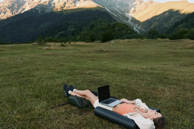 A person rests on a grassy field with a laptop, enjoying a sunny day in nature. The tranquil scene conveys relaxation, creativity, and productivity outdoors.