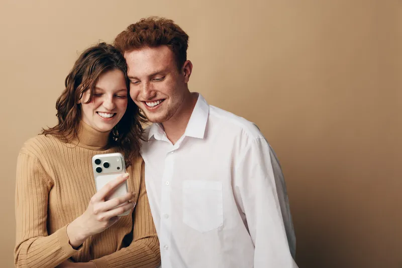 Smiling young couple looks at smartphone screen together, sharing a joyful moment. Casual clothing and warm beige background create a cozy and intimate atmosphere.