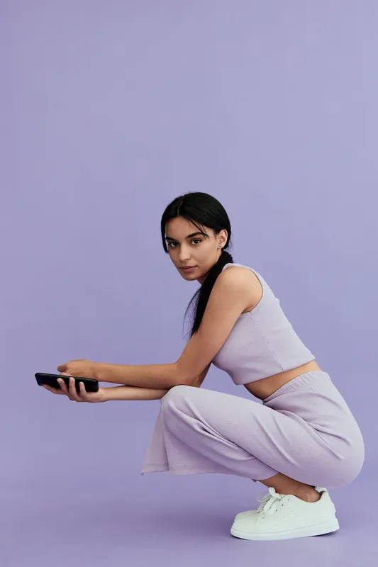 Young woman in lavender outfit crouching on purple background holding remote control, casual style, focused expression, minimalistic setting, soft lighting, sporty shoes.