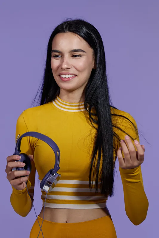 Woman sport fitness holding headphones, dressed in yellow activewear, smiling with long dark hair against purple background in a studio setting.