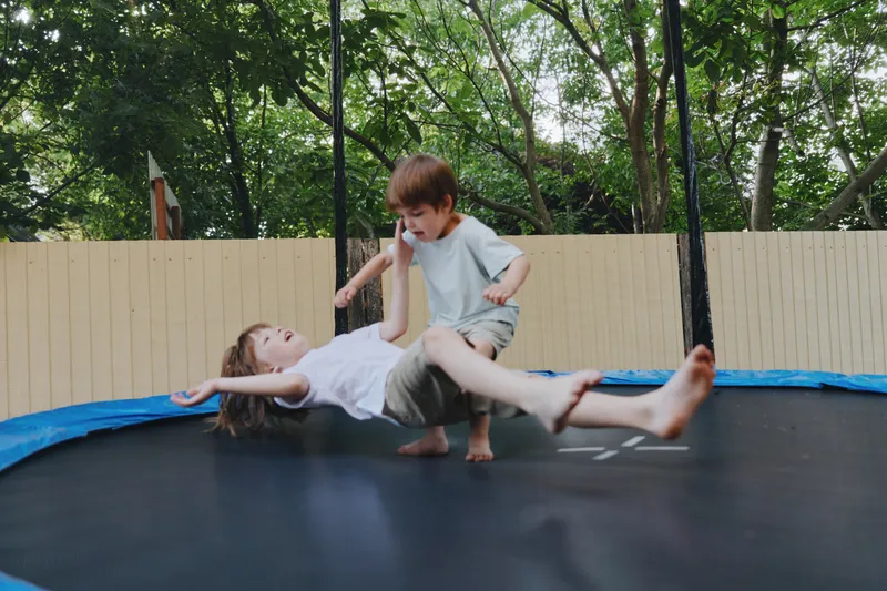 Two playful kids jumping on a trampoline, enjoying a fun moment in the garden, with bright sunlight filtering through lush green trees around them.
