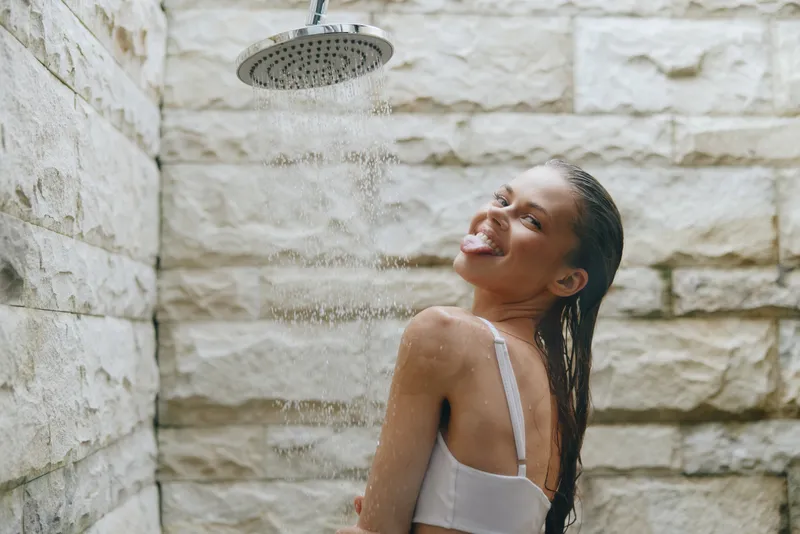 smiling woman enjoying outdoor shower under rain with wet hair by stone wall, carefree and playful moment in natural light