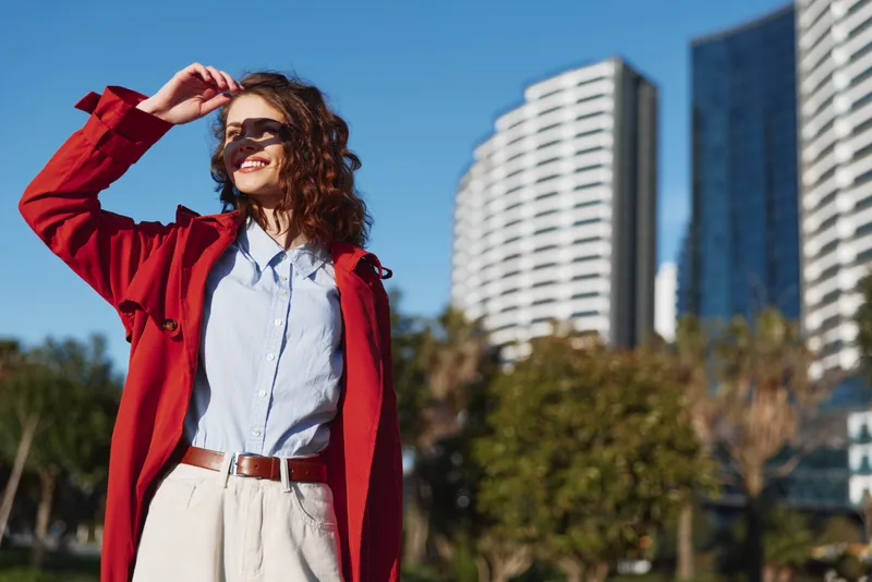 Smiling woman wearing a red coat and light blue shirt stands outdoors in an urban setting, captured in bright daylight for a vibrant fashion and lifestyle mood.