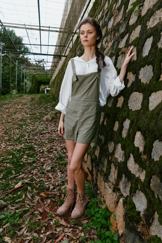 Young woman in a casual green dress stands confidently against a mossy stone wall in a serene outdoor setting, embodying a peaceful and natural vibe