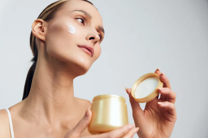 Young woman applying cream on face, holding open jar, skincare routine, clean skin, soft lighting, white background, focused expression, beauty care concept