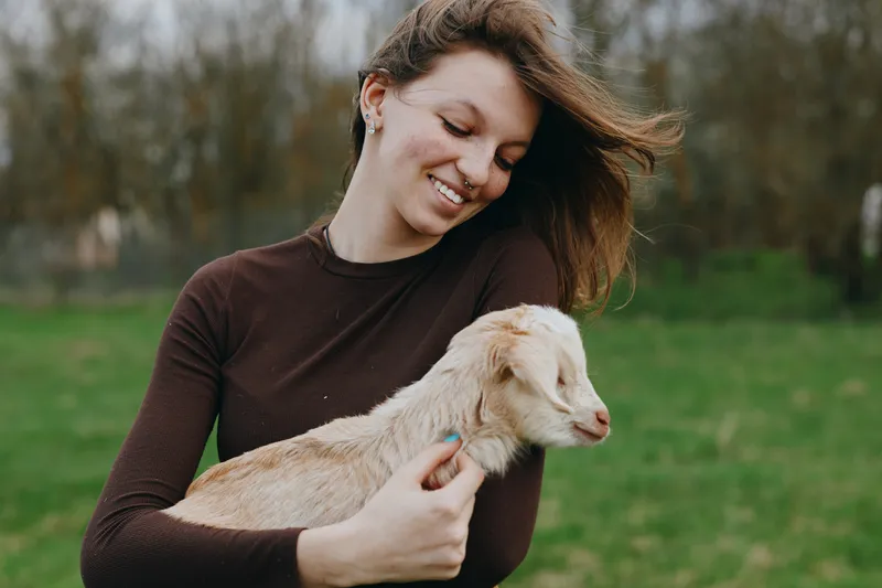 Lifestyle moment outdoors featuring a smiling woman holding a gentle goat kid, sunlit meadow, soft breeze, warm tones, candid connection, serene mood, natural colors, authentic countryside ambiance
