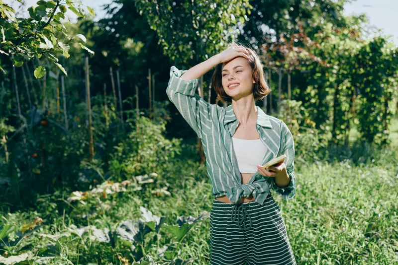 Young woman in a sunlit garden shields her eyes with one hand, wearing a green striped shirt and white top, while holding a book.