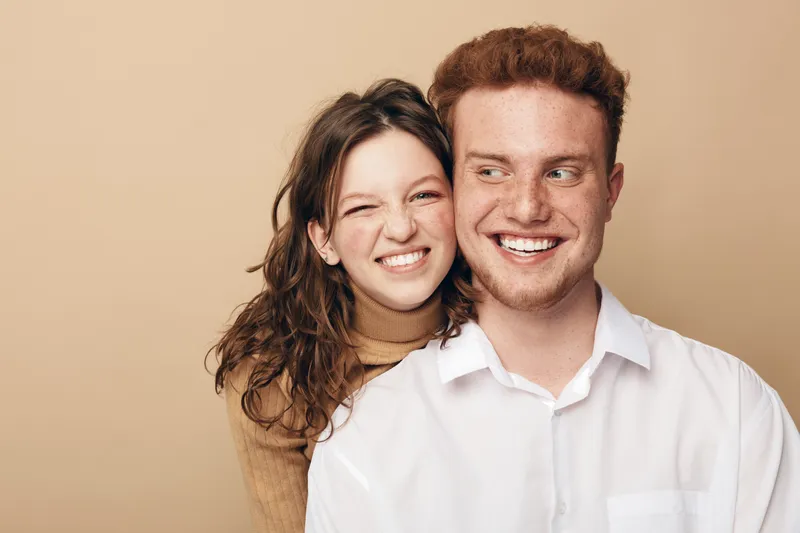Happy young couple with smiling faces posing together on beige background. The woman embraces the man from behind, both showing joyful expressions and casual style clothing.