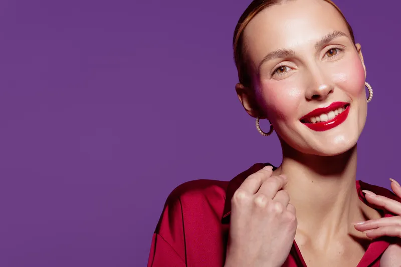 Smiling woman with red lipstick and hoop earrings poses against purple background. She wears a red top and shows confident, joyful expression. Concept of beauty, fashion, and makeup.