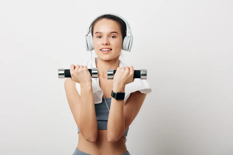 Woman fitness enthusiast lifting dumbbells with headphones in a bright studio, showcasing workout strength, focus, and sport performance during training.