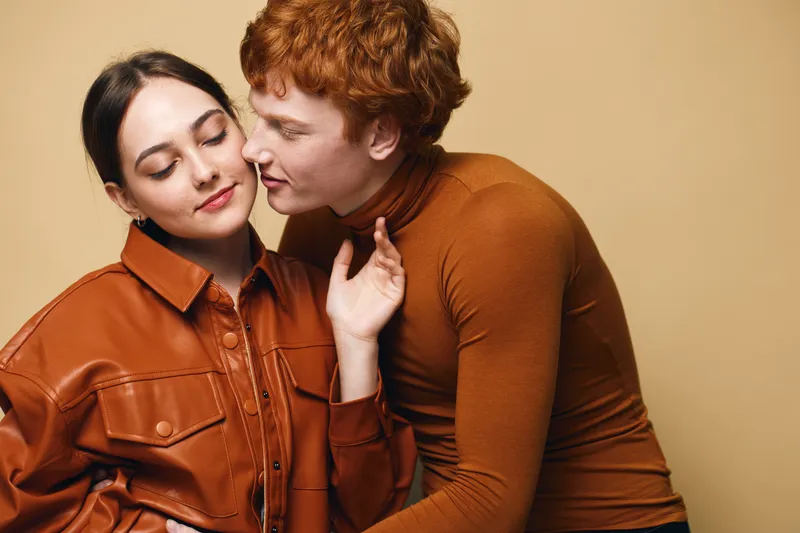 Couple in love with woman wearing brown leather jacket and man in brown turtleneck posing on beige background. Closeup romance and affection between young people.