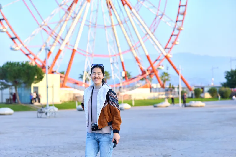 Woman in casual jacket smiles while walking beside a large ferris wheel at an outdoor amusement park, capturing a lively scene of leisure, sunshine, and urban play