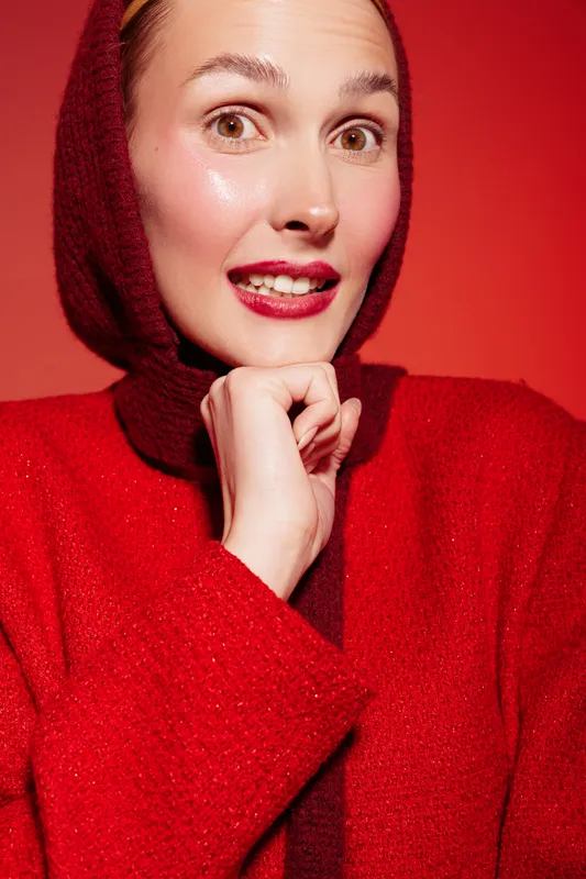 Smiling woman in red sweater and hood poses with hand near chin on red background. Bright makeup and natural skin glow create cheerful and confident mood in studio portrait photography.