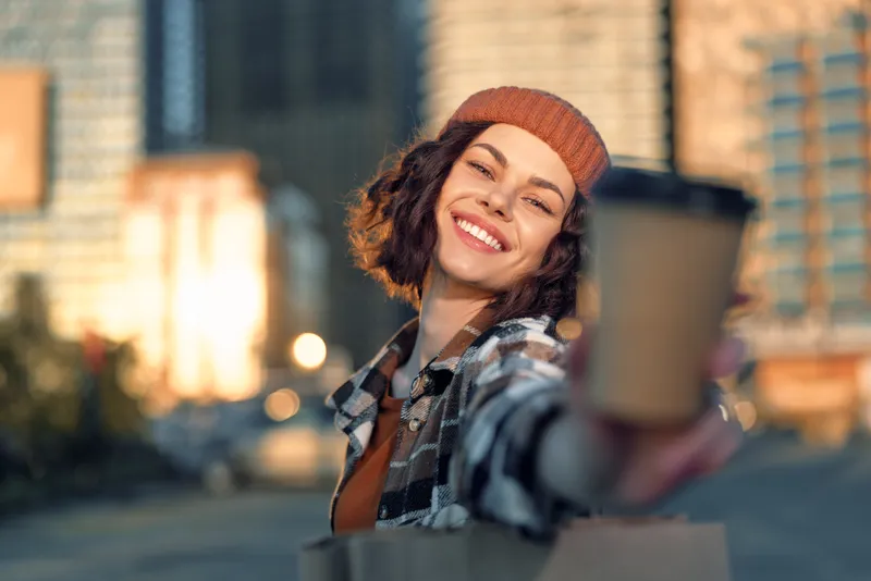 woman smile coffee urban candid lifestyle portrait at golden hour offering takeaway cup and shopping bag, authenticity and emotional storytelling with mindful living and warm golden hour glow