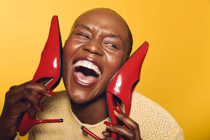 Happy African American woman with dark skin laughing joyfully holding red high heels near face on yellow background. Concept of fashion, beauty, confidence, and vibrant lifestyle.