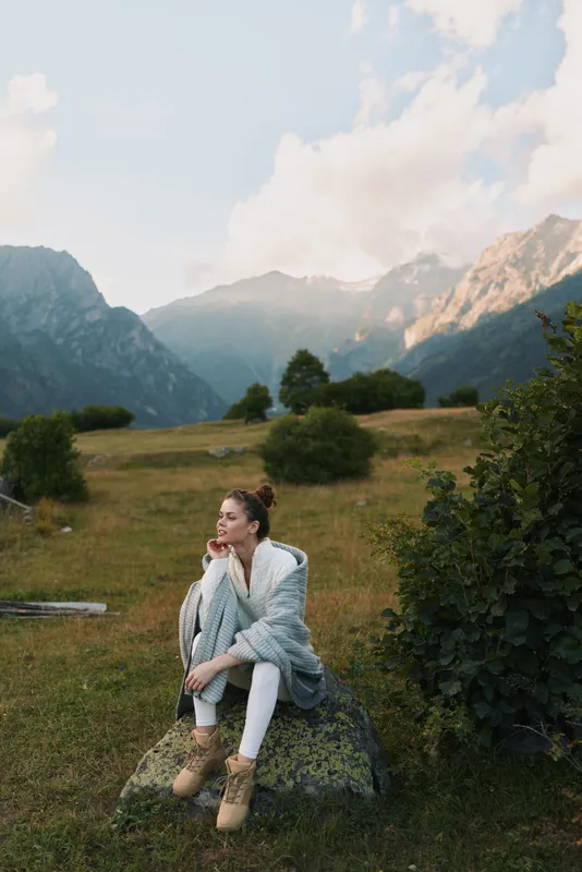 Woman sitting on a rock in a mountainous meadow, enjoying the outdoors in a cozy cardigan. Calm nature scene conveys lifestyle and peaceful contemplative fashion mood.