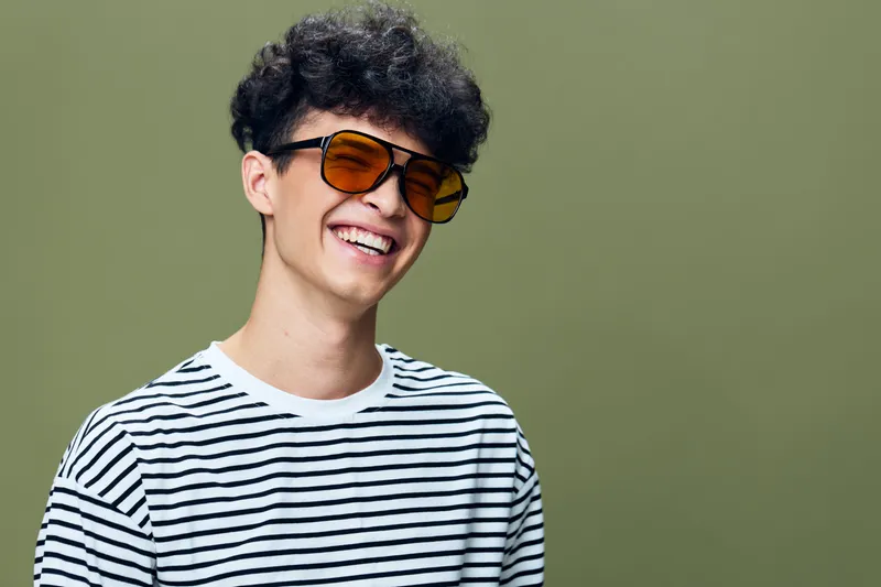 smiling man with curly hair wearing striped t-shirt and orange sunglasses on green background, cheerful expression, casual style, youthful energy