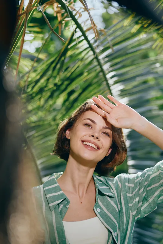 Woman, outdoors, smiling portrait in natural light as she stands among lush green leaves, relaxed pose and warm expression, sunny mood in a tropical garden setting, vibrant colors and fresh air.