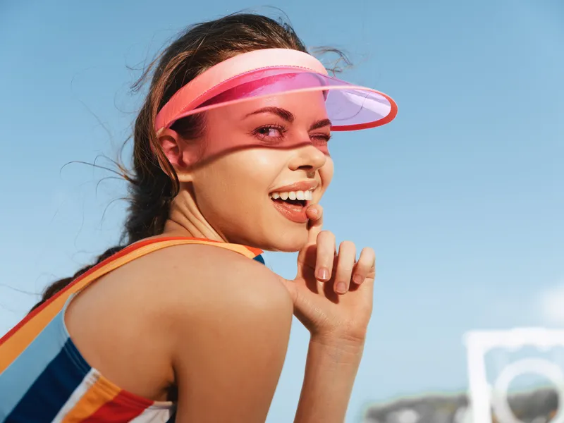 Cheerful young woman wearing a colorful sleeveless top and a pink visor smiles brightly against a clear blue sky, capturing a vibrant outdoor leisure moment.