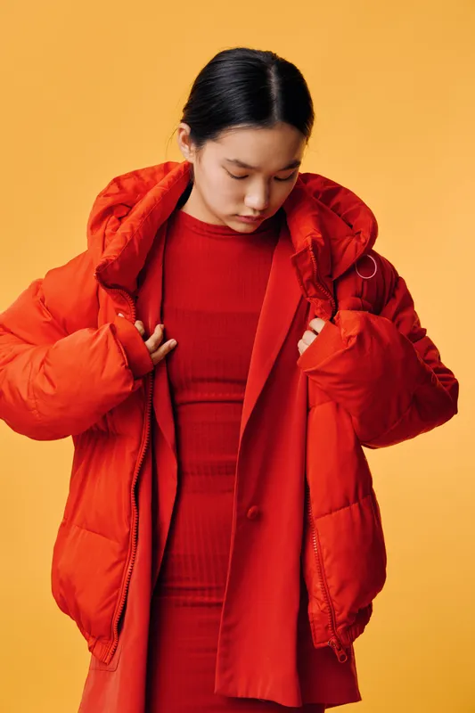 Young woman wearing red oversized puffer jacket and red outfit looking down on yellow background. Fashion style with winter coat and casual apparel in studio setting with thoughtful pose.