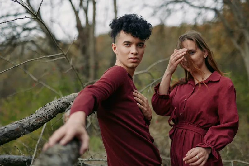 A natural portrait of a young couple in matching burgundy outfits in a quiet woodland, conveying authenticity and credibility through relaxed poses, direct gaze, warm skin tones and textured fabric.
