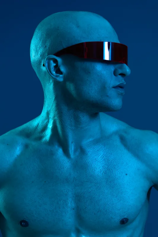 Portrait of a man with a shaved head and futuristic sunglasses, exuding a confident look. The blue light lends a striking atmosphere to the black and white image.