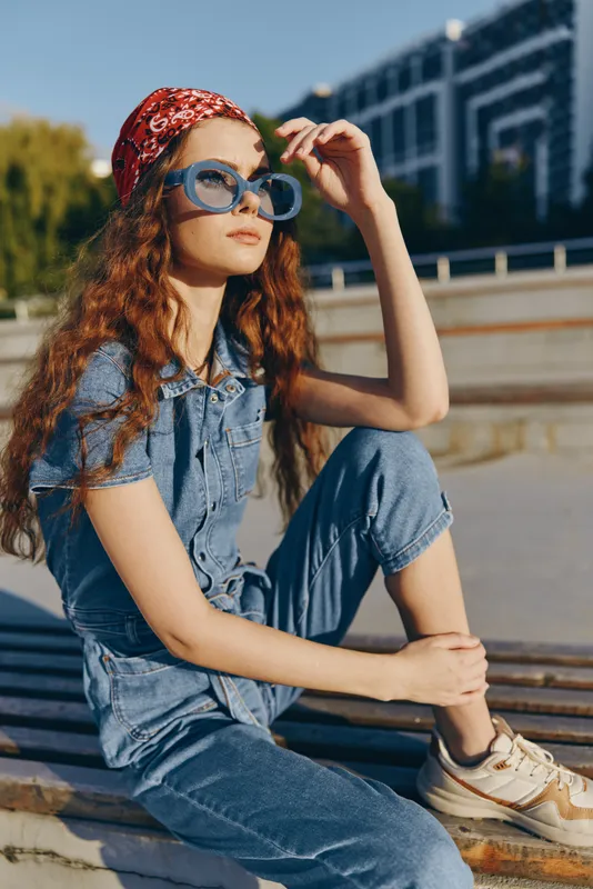 Lifestyle woman in modern boho-western denim street style wearing blue sunglasses and a red bandana, captured with warm film color tones under natural sunlight.