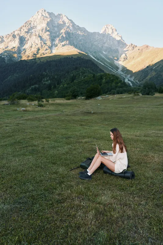 A young woman sits on a grassy field with a laptop, surrounded by towering mountains. She works outdoors, relaxed and focused, enjoying nature and a peaceful atmosphere.