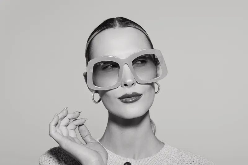 Slim caucasian woman with fair skin and slicked hair wears oversized square glasses and hoop earrings. Studio portrait in black and white with neutral background and confident pose.