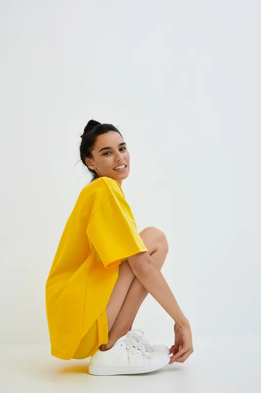 young woman crouching in oversized bright yellow shirt with white sneakers against white background, casual style, minimalistic mood, studio setting