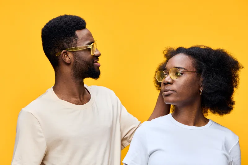 Happy young couple wearing sunglasses, with a vibrant yellow background, showcasing love and connection through playful emotions and stylish fashion