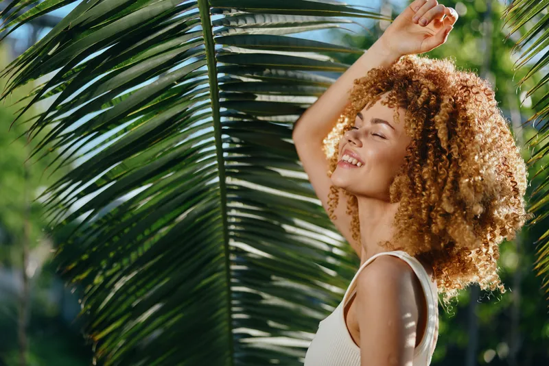 Smiling woman with curly hair enjoys sunny tropical environment surrounded by lush green palm leaves wearing white sleeveless top.