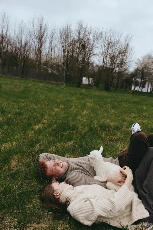 Lifestyle scene shows a couple lying on grass outdoors with a curious goat kid, wearing cozy knitwear, sharing affectionate smiles in a relaxed countryside moment under soft daylight.