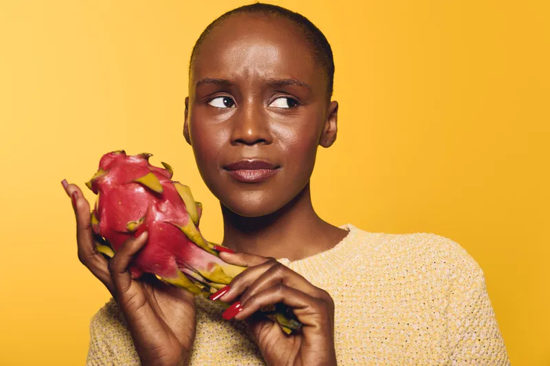 Portrait of a thoughtful dark skinned African woman holding a dragon fruit with both hands against yellow background. Concept of healthy lifestyle, exotic fruit, and natural beauty.