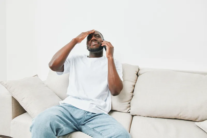 Young African American man with a distressed expression talking on the phone on a couch, wearing casual clothing in a neutral toned room