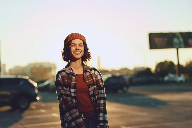Woman smile candid urban beanie lifestyle portrait in a sunlit parking lot at golden hour glow, authenticity and candid lifestyle emphasized, mindful living captured with emotional storytelling.