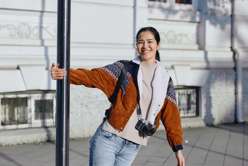 Woman leans on a street pole in a casual autumn outfit, orange brown jacket and cream sweater, denim jeans with crossbody bag, outdoor urban scene, bright mood and natural smile