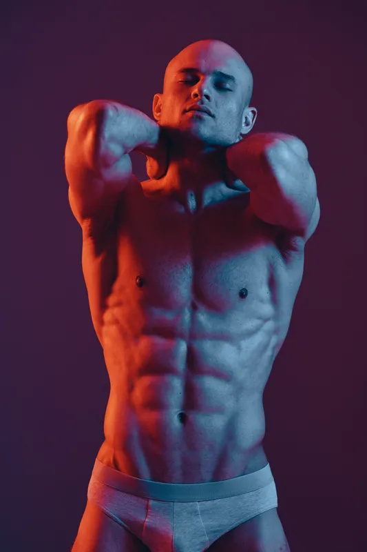 A confident man poses with strong muscles under vibrant lighting, showcasing a dynamic blend of blue and red tones, emphasized by the dramatic shadows and highlights.