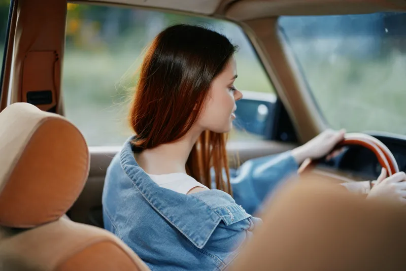 Young woman driving a vintage car, wearing a denim jacket, showcasing a relaxed vibe while enjoying a scenic journey with a natural background.