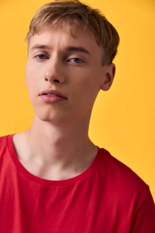 Close up of a young man wearing a red shirt against a bright yellow background, neutral expression, studio lighting, fashion and lifestyle oriented visual for marketing and editorial use