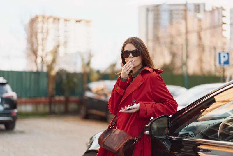 Cheerful woman outdoors in a bright red coat beside a car on a city street, sunglasses on, candid moment, soft afternoon light, urban fashion vibe, confident pose today