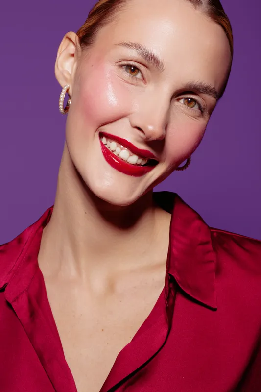 Smiling woman with red lipstick and shiny skin wears red blouse and hoop earrings. Close-up portrait on purple background. Concept of beauty, makeup, fashion, and confident expression.