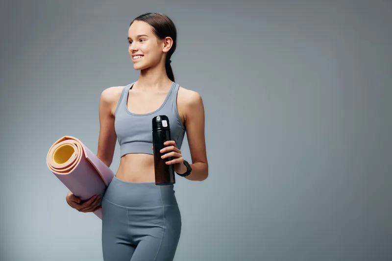 Fitness woman in athletic wear holding a yoga mat and water bottle, ready for training or gym session, bright studio background and a healthy lifestyle vibe.