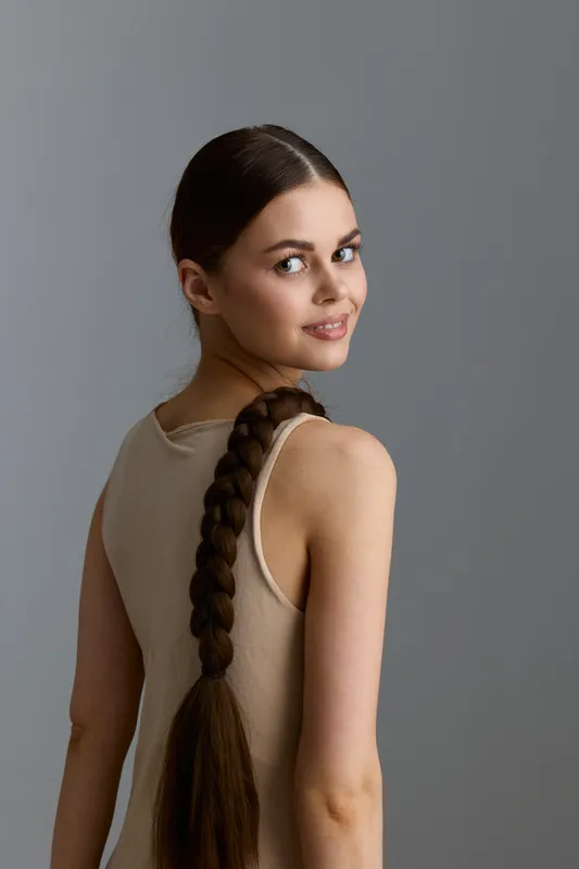 young woman with long braided hair wearing a beige sleeveless top smiles over shoulder against plain gray background, natural light highlights her clear skin and warm expression.