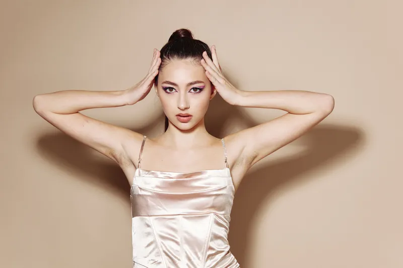 Elegant young woman with clear skin and slicked hair poses with hands on head in a soft satin dress against neutral background. Concept focuses on beauty fashion and creative style.