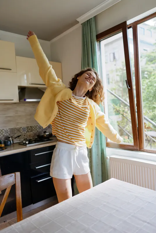 Young woman dances in a bright kitchen, arms raised in joy, smiling broadly as sunlight pours through the window, capturing a candid cheerful lifestyle moment