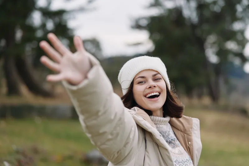 Woman in a winter coat and beanie smiles while reaching toward the camera in an outdoor park, showing joyful energy, natural candid charm and relaxed everyday style.